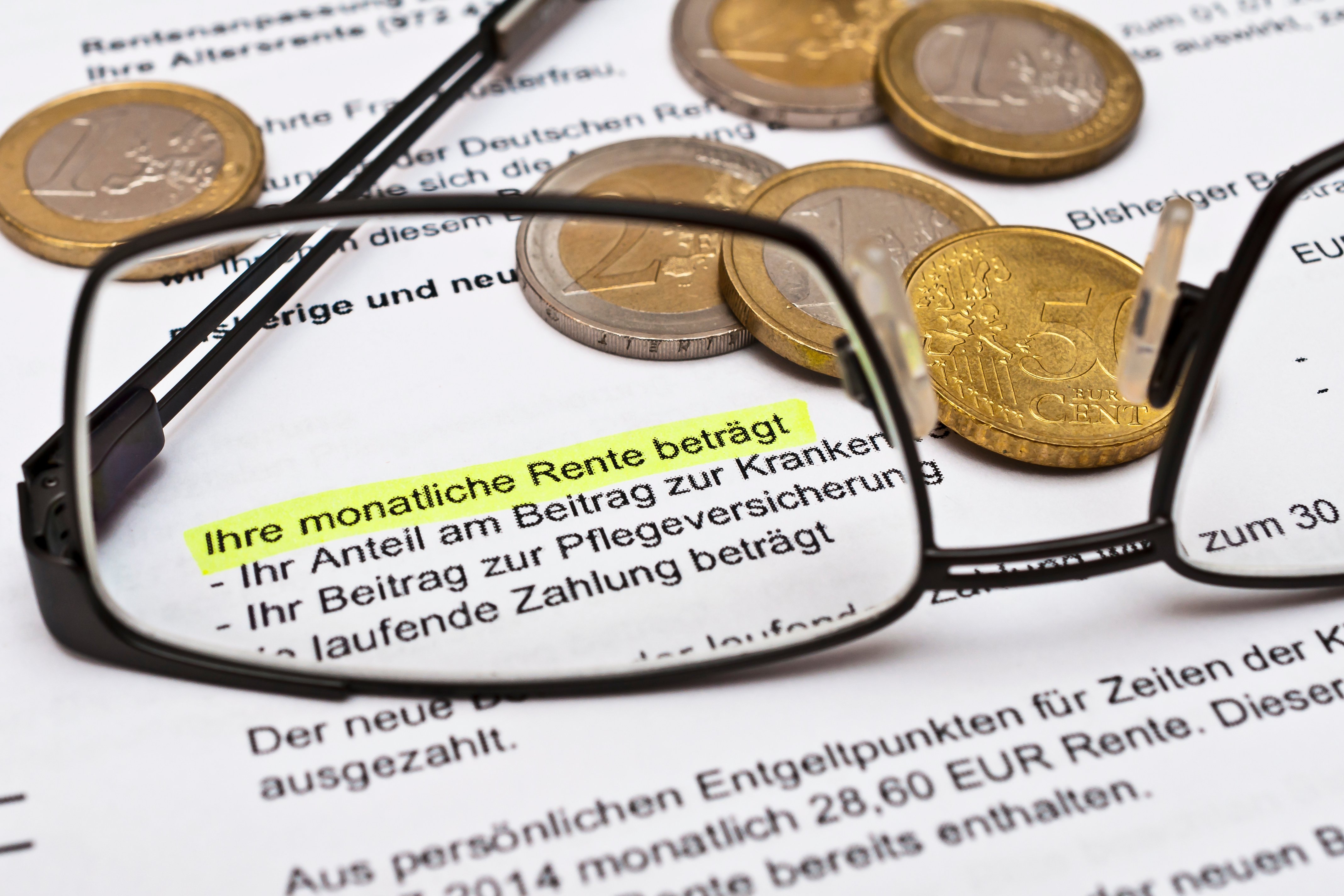 Nahaufnahme eines Rentenbescheids mit markiertem Text ‚Ihre monatliche Rente beträgt‘, umgeben von Euro-Münzen und einer Brille im Vordergrund.