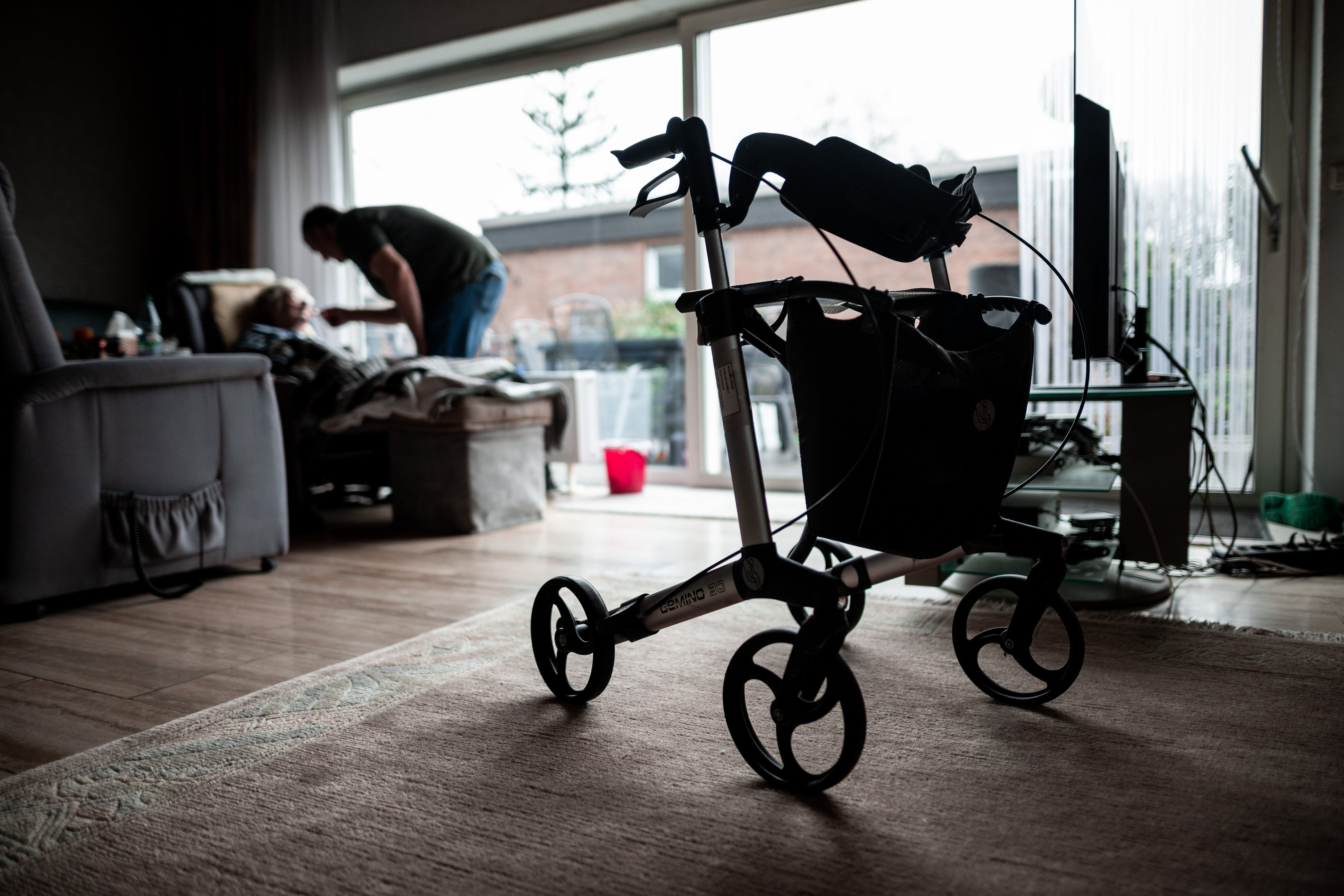 Häusliche Pflege Blick in ein dunkles Wohnzimmer. Im Vordergrund steht ein Rollator. Im Hintergrund sieht man eine sitzende Person, die von einer anderen Person, die sich über sie beugt, gefüttert wird. Symbolfoto für Angehörigenpflege