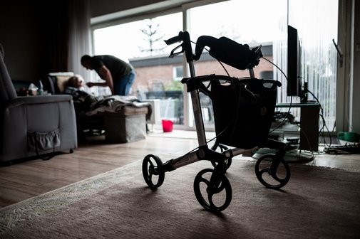 Häusliche Pflege Blick in ein dunkles Wohnzimmer. Im Vordergrund steht ein Rollator. Im Hintergrund sieht man eine sitzende Person, die von einer anderen Person, die sich über sie beugt, gefüttert wird. Symbolfoto für Angehörigenpflege