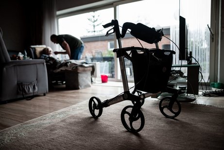Blick in ein dunkles Wohnzimmer. Im Vordergrund steht ein Rollator. Im Hintergrund sieht man eine sitzende Person, die von einer anderen Person, die sich &uuml;ber sie beugt, gef&uuml;ttert wird. Symbolfoto f&uuml;r Angeh&ouml;rigenpflege