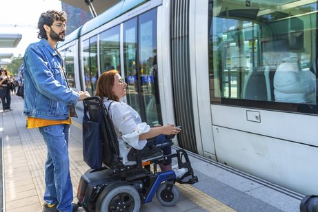 Barrierefreiheit im ÖPNV Junger Mann, der einer erwachsenen Frau im Rollstuhl beim Einsteigen in eine moderne Straßenbahn an einer belebten Haltestelle hilft, um Barrierefreiheit und Inklusion zu unterstreichen
