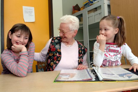 Seniorin mit zwei Kindern, sie üben Lesen, vor ihnen liegt ein Ordner mit Arbeitsblättern.
