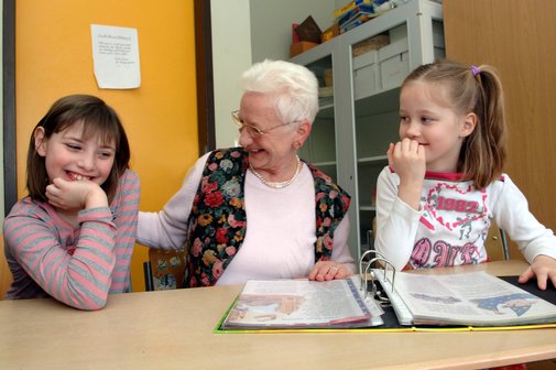 Seniorin mit zwei Kindern, sie üben Lesen, vor ihnen liegt ein Ordner mit Arbeitsblättern.