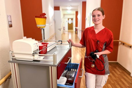 Michelle Pflegefachkraft Das Foto zeigt Michelle. Sie steht neben einem Medikamentenwagen im Flur des Pflegeheimes und hält ein Blutdruckmessgerät in der Hand.