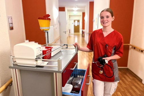Michelle Pflegefachkraft Das Foto zeigt Michelle. Sie steht neben einem Medikamentenwagen im Flur des Pflegeheimes und hält ein Blutdruckmessgerät in der Hand.
