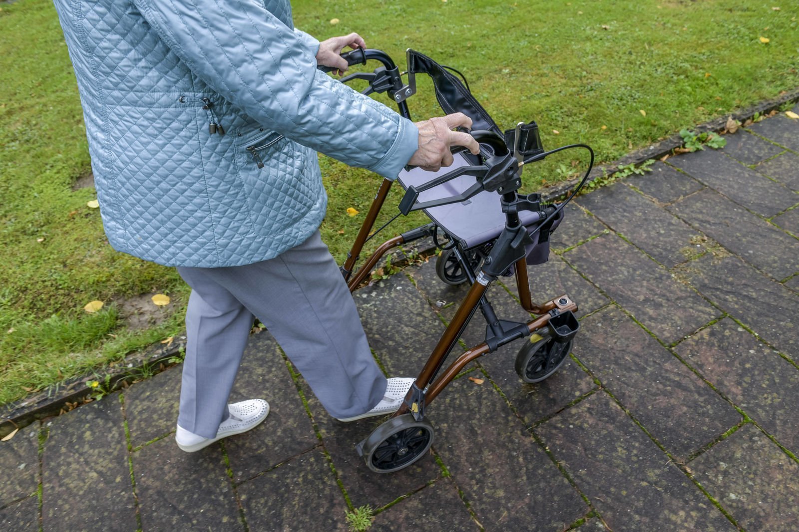 Eine alte Dame geht mithilfe eines Rollators draußen auf einem gepflasterten Weg.