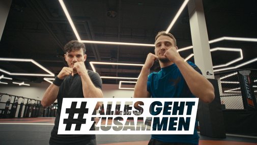 Zwei junge Männer in einer Sporthalle, sie haben bei die Fäuste in einer Boxer-Pose gehoben, blicken frontal in die Kamera. Darüber das Logo der Aktion Alles geht zusammen.