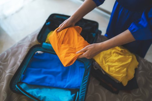 Eine Frau packt Kleidungsstücke in einen kleinen Koffer, der auf einem Bett liegt.