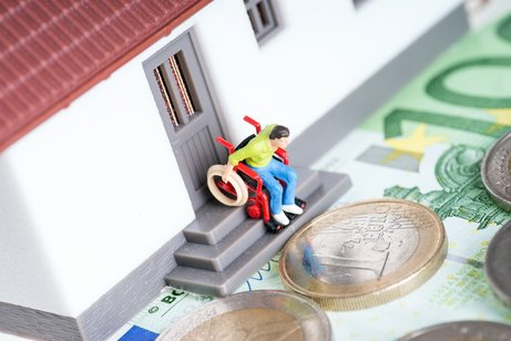 Ein Spielzeughaus, auf der T&uuml;rschwelle sind Stufen. Ein Fig&uuml;rchen im Rollstuhl steht an der Schwelle, schaut nach unten. Vor dem Spielzeughaus liegen Geldscheine und -m&uuml;nzen.