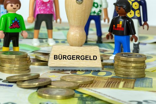 Symbolbild: Stempel mit der Aufschrift Bürgergeld und Spielfiguren auf Euro-Banknotenoten