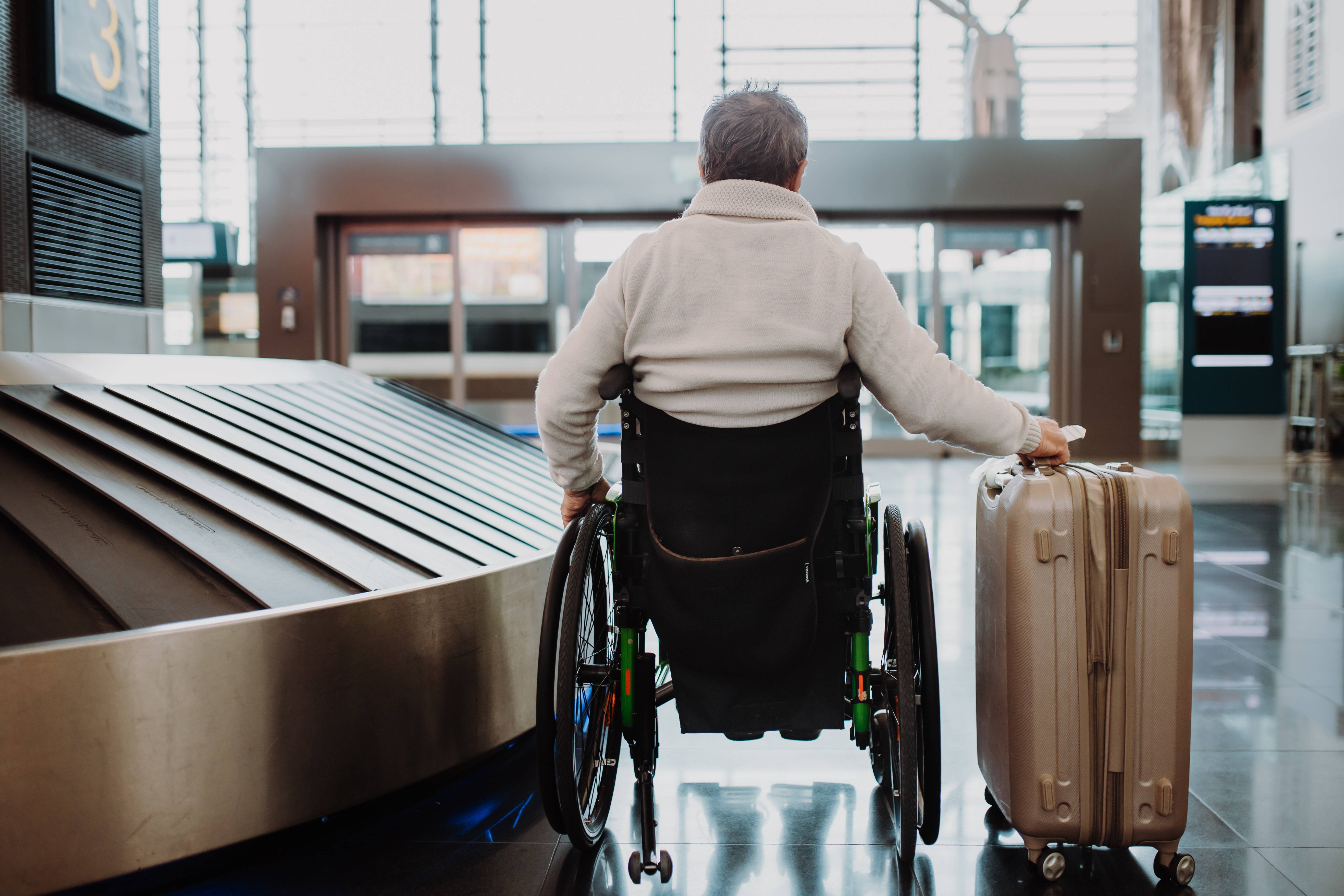 Passagier mit Behinderung am Flughafen Rückenansicht eines älteren Mannes im Rollstuhl, der am Gepäckband eines Flughafens steht. Neben ihm steht ein Reisekoffer.