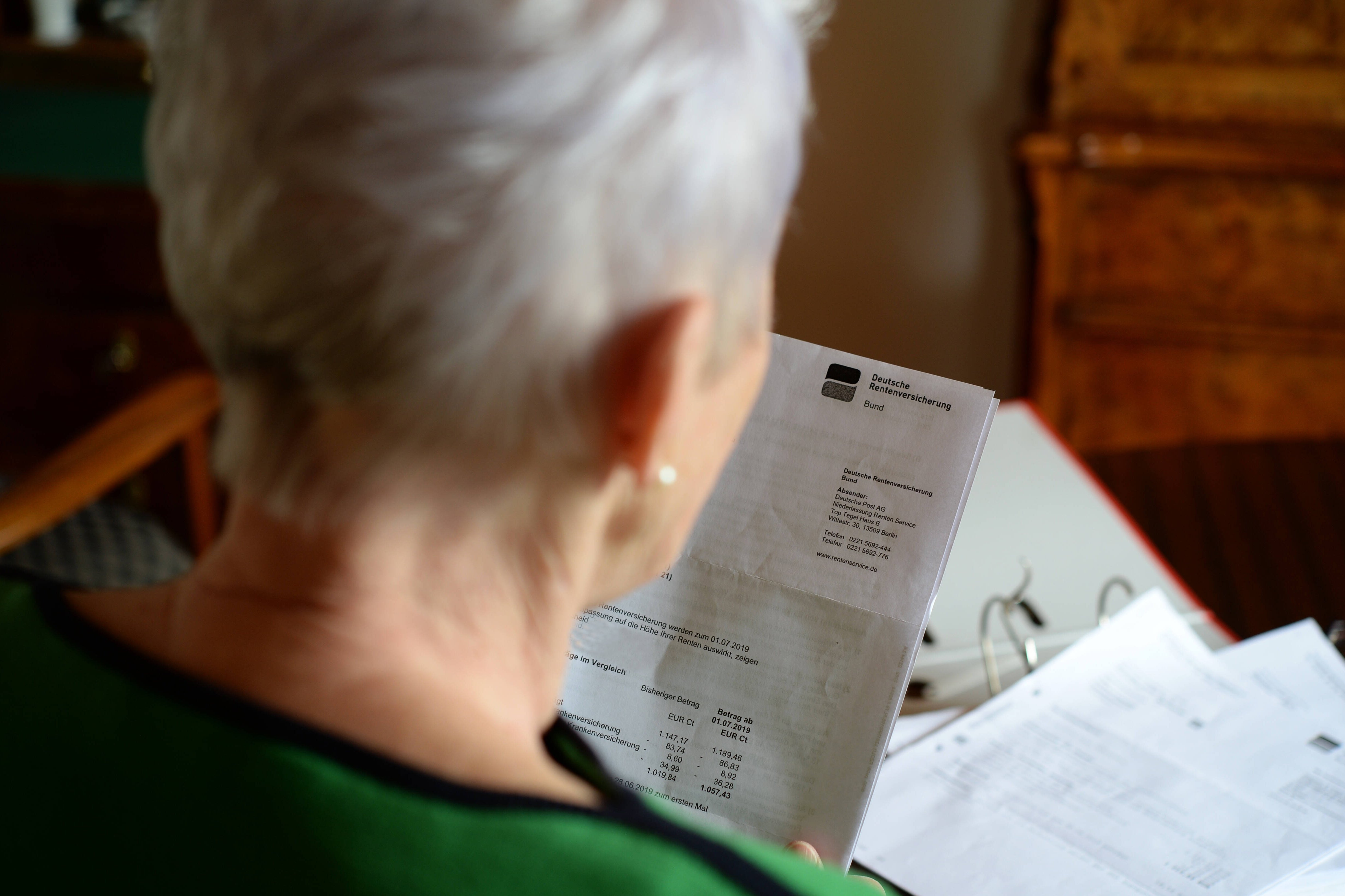 Rentenerhöhung Blick über die Schulter einer älteren Frau, sie hat einen Brief der Deutschen Rentenversicherung und einen Ordner mit Unterlagen vor sich liegen.