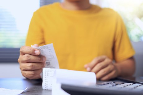 Eine Person im gelben Pullover sitzt an einem Schreibtisch, vor sich einen Taschenrechner und ein Dokument, wahrscheinlich eine Rechnung, als Symbol f&uuml;r steigende Kosten