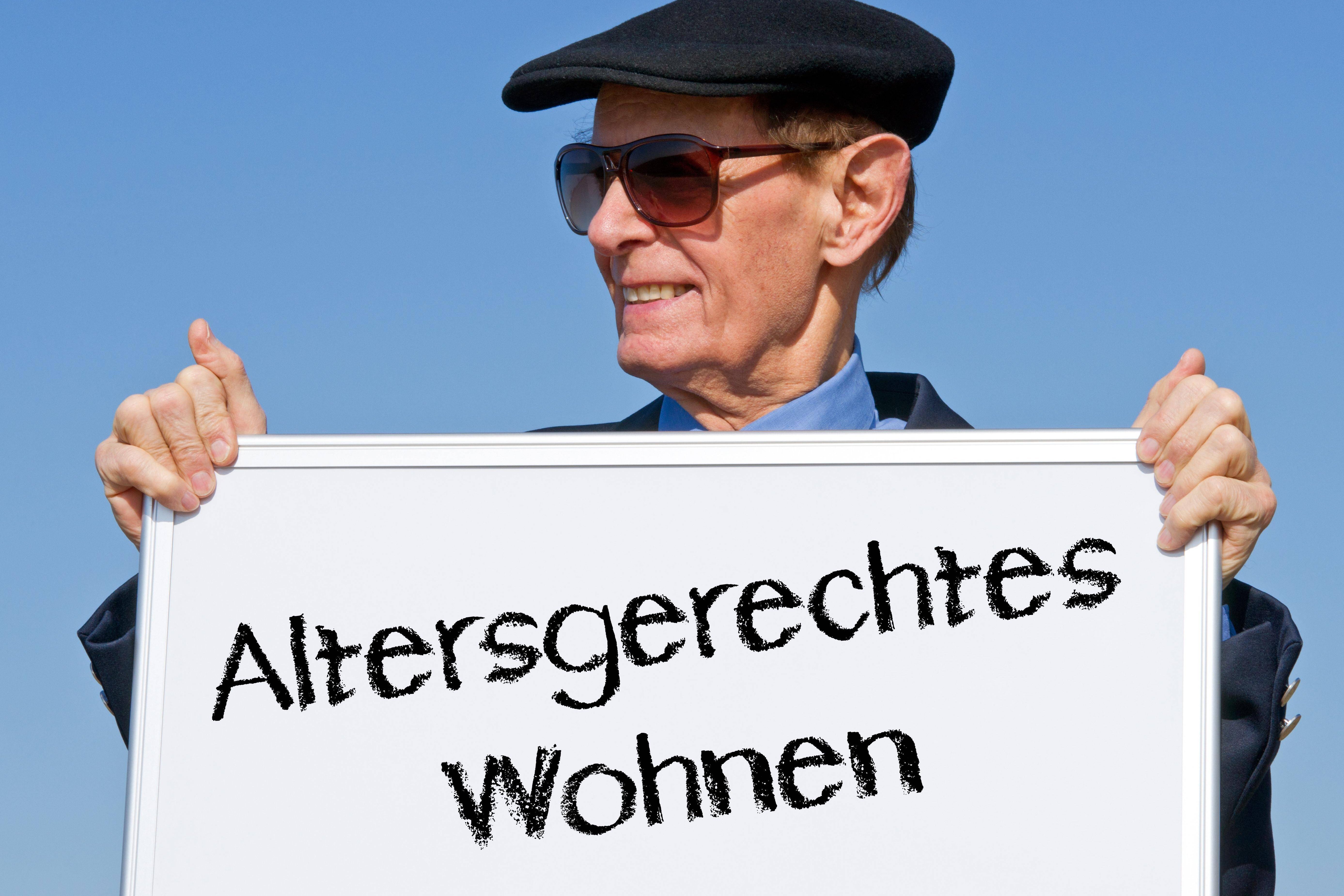 Altersgerechtes Wohnen Senior mit Schirmmütze und Sonnenbrille hält ein Schild in beiden Händen, auf dem steht "Altersgerechtes Wohnen"