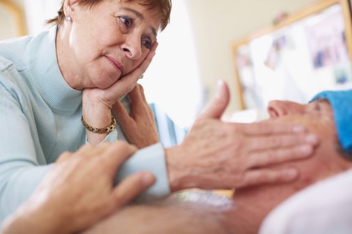 Eine ältere Frau beugt sich über ihren pflegebedürftigen Mann, legt ihm liebevoll eine Hand auf die Stirn. Sie sieht besorgt aus. 