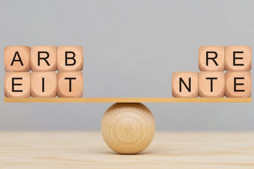 Holzbuchstaben werden auf einer Wippe aus einer Holzkugel und einem kleinen Holzbrett ausbalanciert. Auf der linken Seite bilden die Buchstabenwürfel das Wort "Arbeit", auf der rechten Seite "Rente".