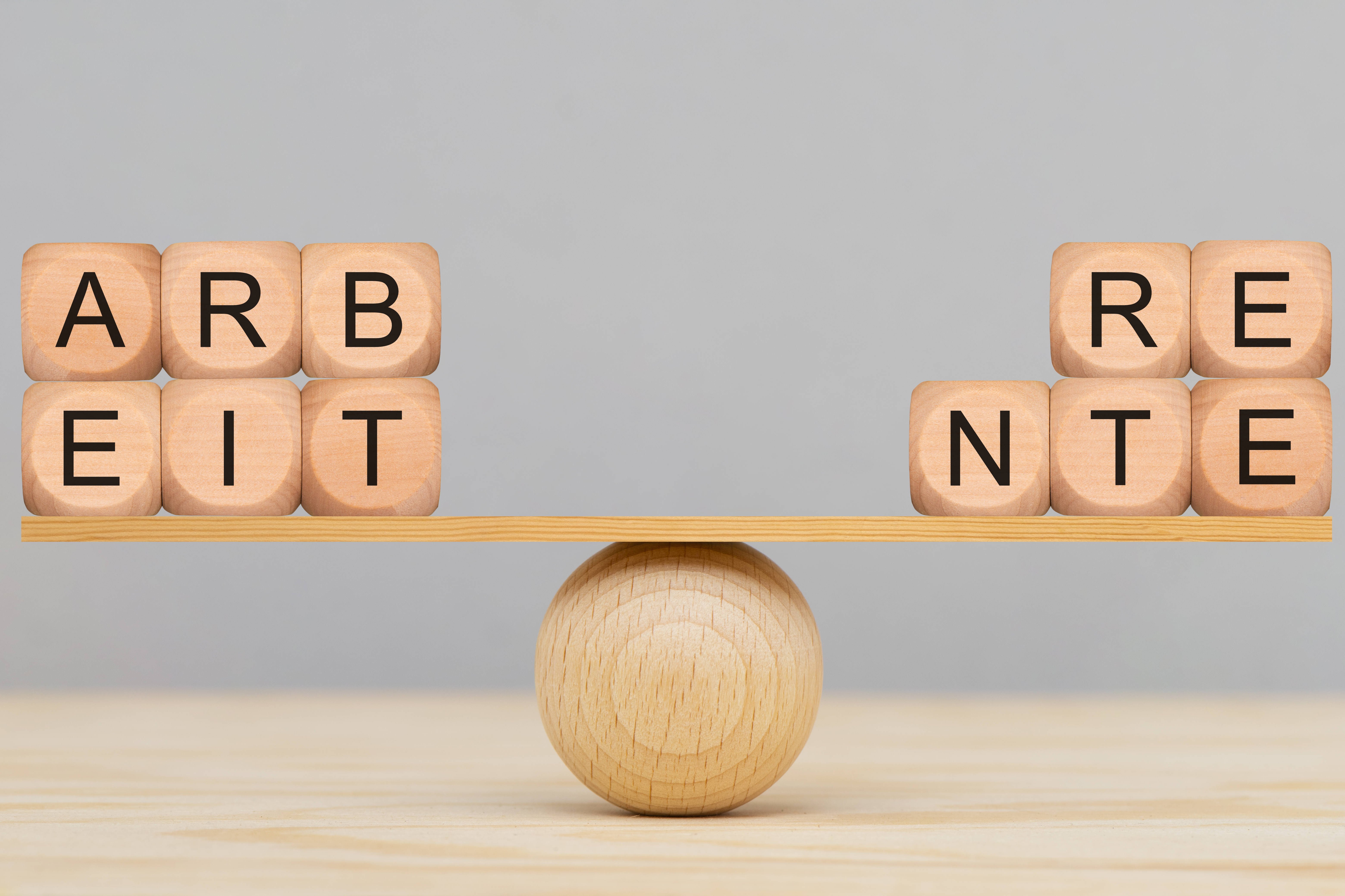 Arbeiten und Rente Holzbuchstaben werden auf einer Wippe aus einer Holzkugel und einem kleinen Holzbrett ausbalanciert. Auf der linken Seite bilden die Buchstabenwürfel das Wort "Arbeit", auf der rechten Seite "Rente".