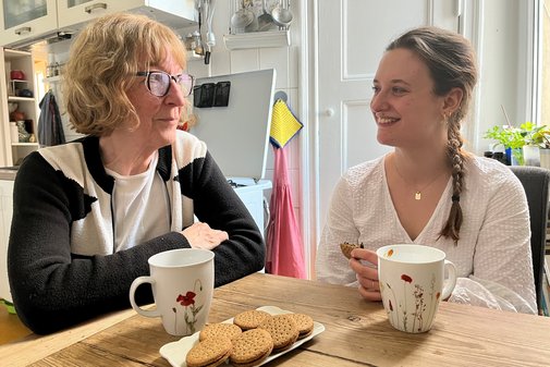 Generationen-WG Cornelia Stauß und Beate Meißner sitzen an einem Holztisch, auf dem Kaffeebecher und ein Teller mit Keksen stehen.