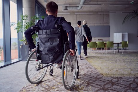 Arbeitnehmer mit Behinderung Rückenansicht eines Mannes im Rollstuhl, der sich durch ein Bürogebäude bewegt, man sieht im Hintergrund andere Mitarbeitende, die nicht im Rollstuhl sitzen.