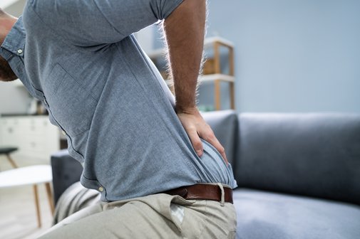 Ein Mann steht vom Sofa auf, hält sich mit einer Hand den unteren Rücken als Symbol für Rückenschmerzen
