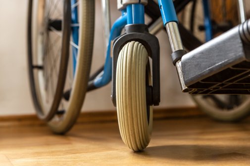 Nahaufnahme eines Rollstuhls: Man sieht ein großes und ein kleines Rad sowie einen Teil des Fußteils