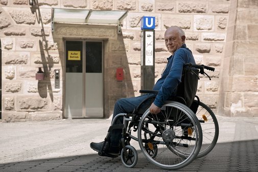 Barrieren im ÖPNV Ein Senior im Rollstuhl steht am unzugänglichen Eingang einer U-Bahn-Station. Dort ist ein Aufzug mit dem Hinweis "Außer Betrieb".