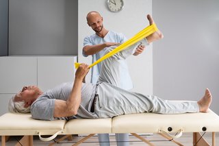 Ein Physiotherapie leitet einen älteren Mann bei einer Übung für das Knie an. Der ältere Mann liegt auf einer Liege und führt eine Knie-Übung mit einem gelben Theraband durch. 