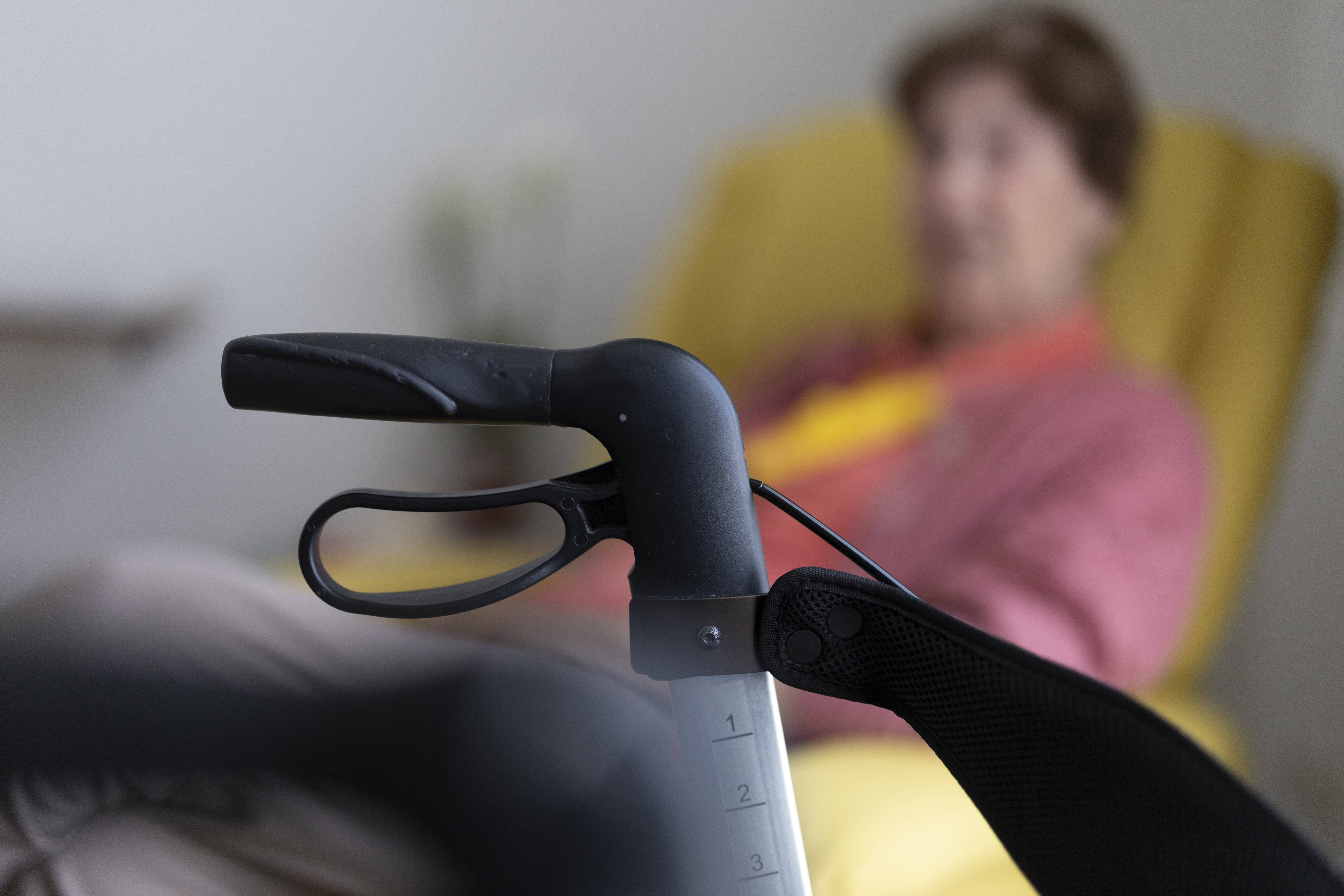 Frau mit Rollator Seniorin sitzt in ihrem Zimmer im Seniorenheim hinter einem Rollator in einem gelben Sessel. Der Rollator im Bildvordergrund ist scharf zu sehen, die Seniorin im Hintergrund verschwommen.