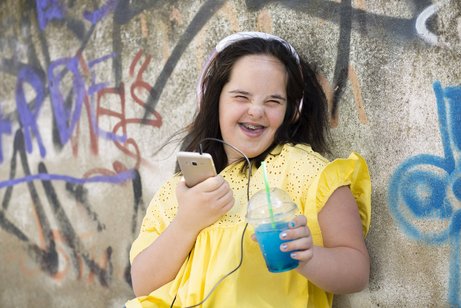 Junge Frau mit Downsyndrom mit ihrem Smartphone Junge Frau mit Downsyndrom, sie lacht in die Kamera, hält ihr Smartphone in der einen Hand und einen Plastikbecher mit einem Getränk in der anderen. Sie trägt Kopfhörer.