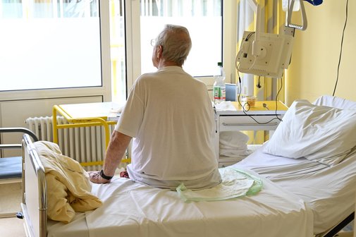 Symbolbild: Ein älterer Patient sitzt in seinem Krankenhauszimmer auf seinem Bett, man sieht ihn in der Rückenansicht