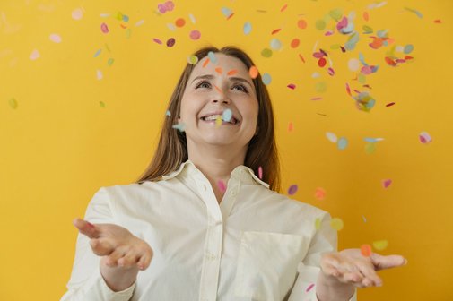 Glückwünsche zum Geburtstag Eine Frau in weißer Bluse fängt lachend buntes Konfetti auf, sie steht vor einem gelben Hintergrund.