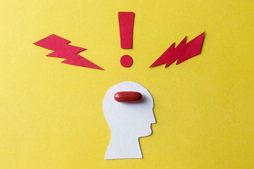 Symbol für Migräne und Kopfschmerzen: Ein aus Pappe ausgeschnittener stilisierter Kopf auf gelben Hintergrund, darin liegt eine rote Pille. Um den Kopf herum symbolisieren zwei rote Blitze und ein rotes Ausrufezeichen Schmerzen.