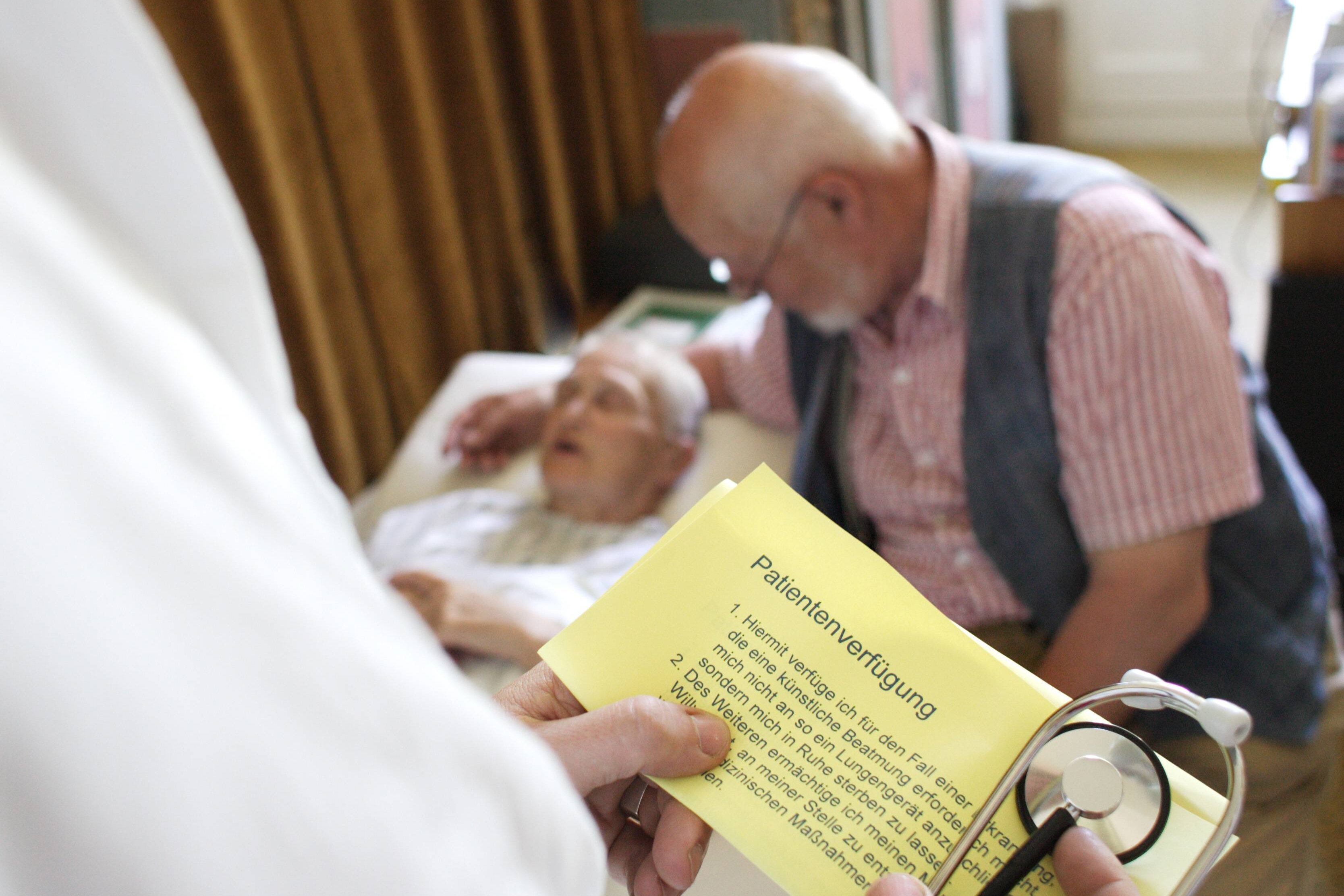 Eine alte Frau liegt im Bett, ein Arzt hält ihre Patientenverfügung in der Hand. 