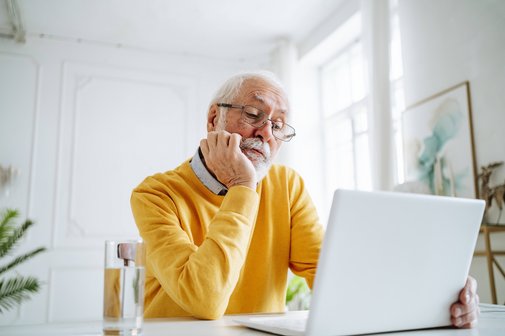 Arbeiten nach der Rente Ein Senior mit Brille im gelben Pullover sitzt an einem Tisch und schaut auf ein Notebook. Das Kinn hat er in die Hand gestützt.