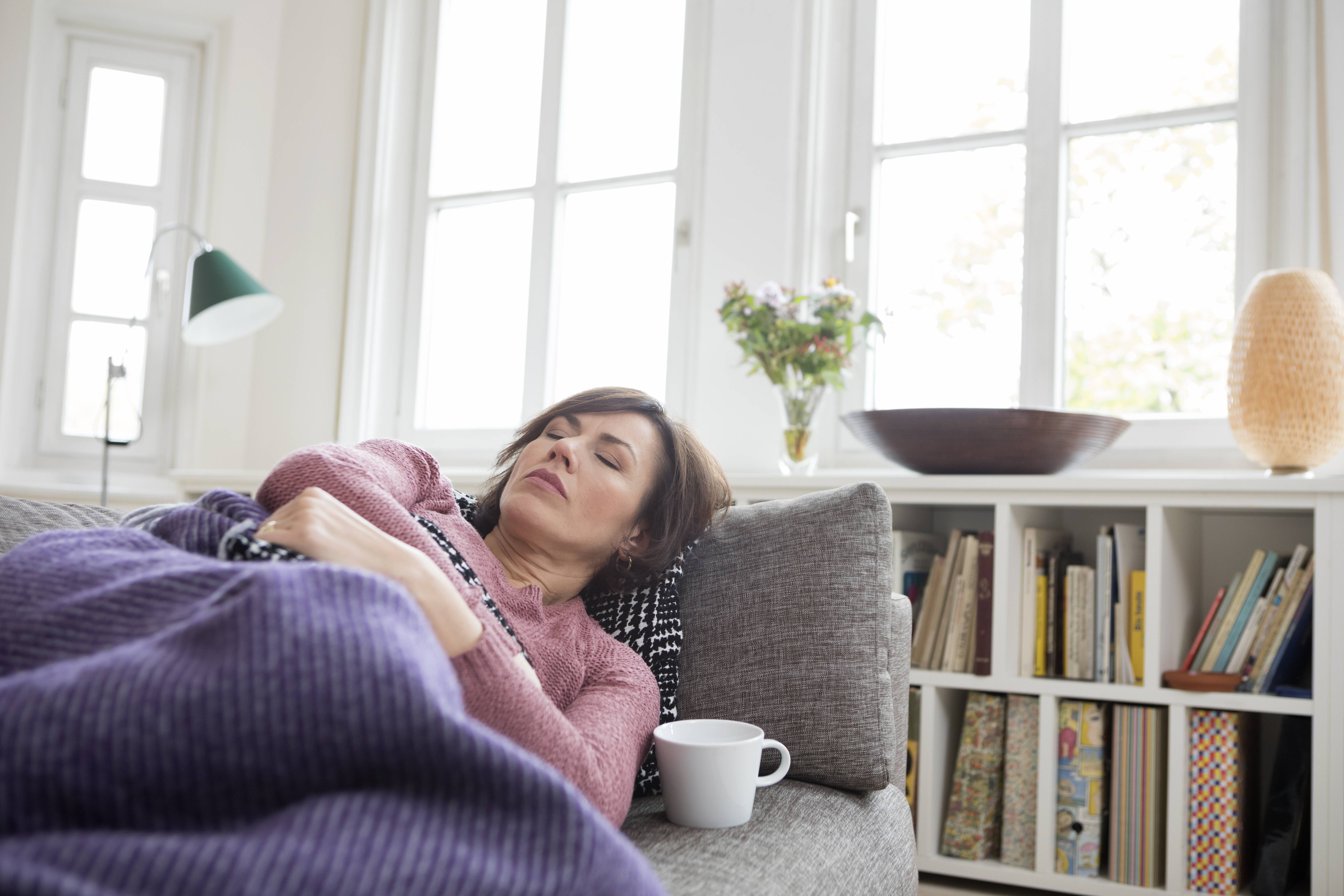 Eine Frau liegt erschöpft auf einer Couch unter einer Decke. 