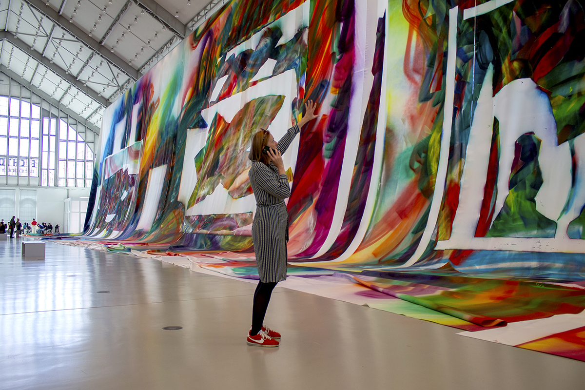 Stefanie Reimers steht in der Ausstellungshalle, betrachtet ein großformatiges Bild und spricht ins Telefon.