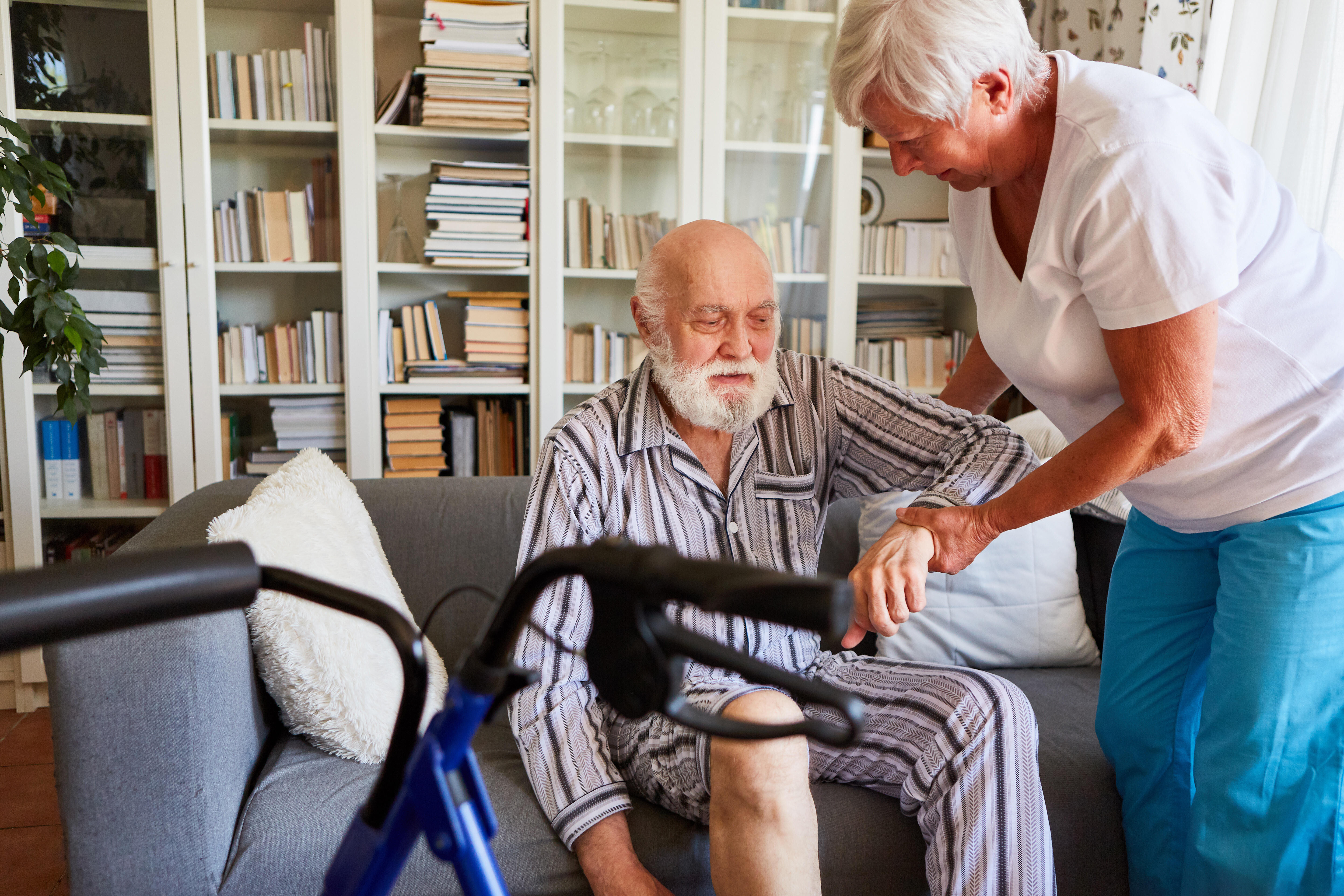 Ein &auml;lterer Mann im gestreiften Pyjama sitzt auf einem Sofa. Eine &auml;ltere Frau h&auml;lt ihn am Arm und hilft ihm aufzustehen. Im Vordergrund steht ein Rollator. 
