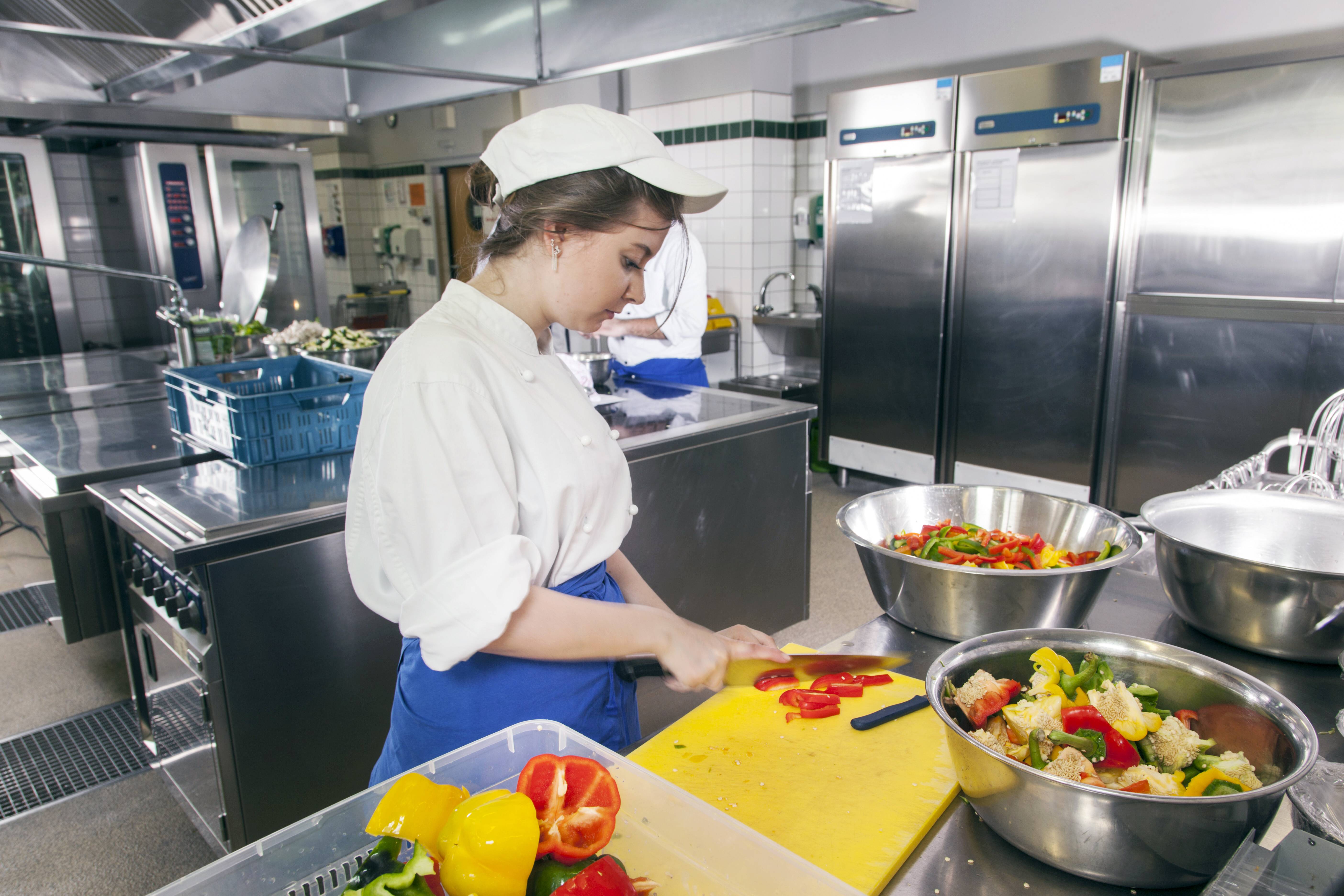 Eine junge Frau in Arbeitskleidung arbeitet in einer Großküche, sie schneidet Tomaten
