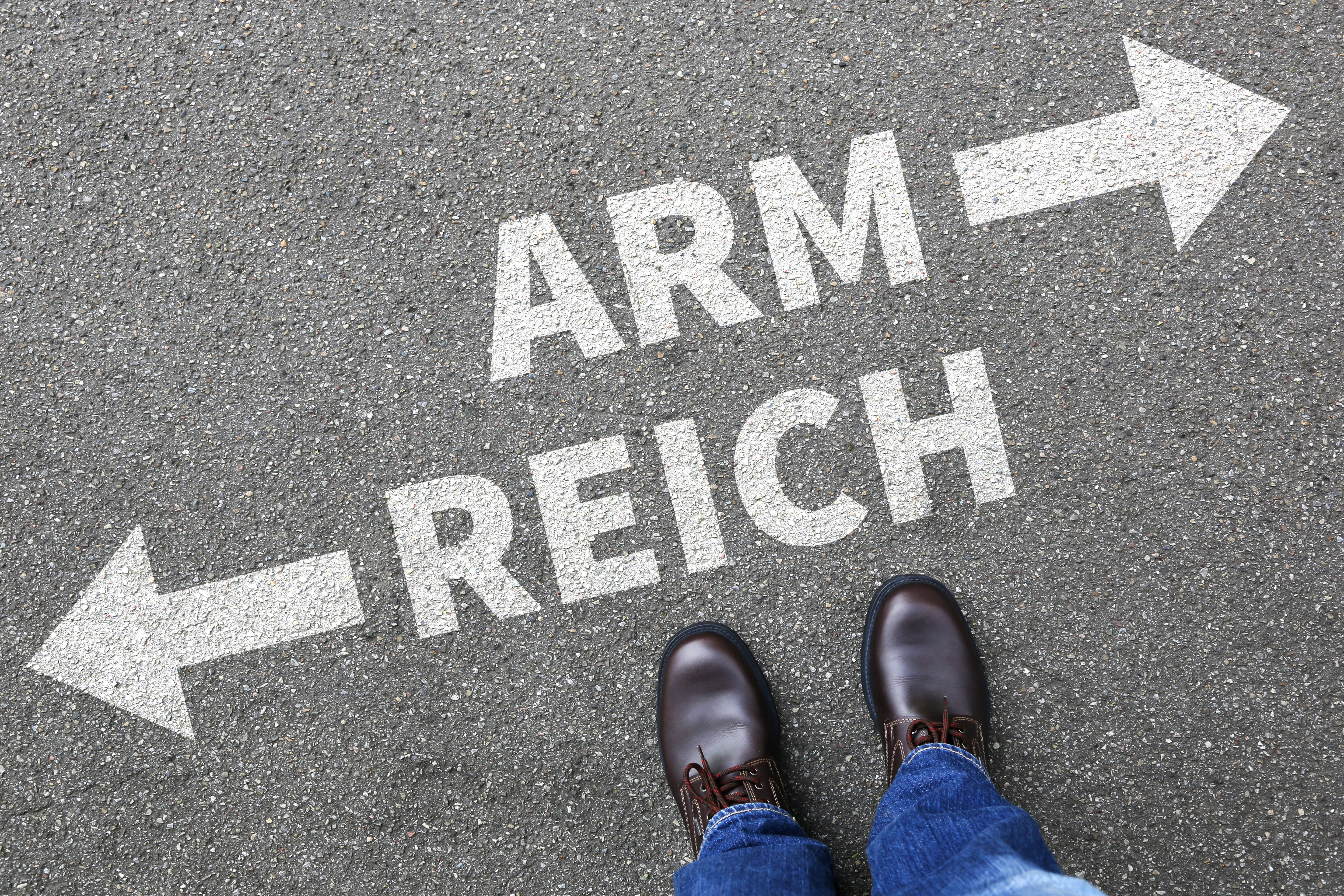 Arm und Reich Füße auf Asphalt, darauf steht in weißer Schrift aufgesprüht "Arm" mit einem Pfeil nach rechts und "Reich" mit einem Pfeil nach links