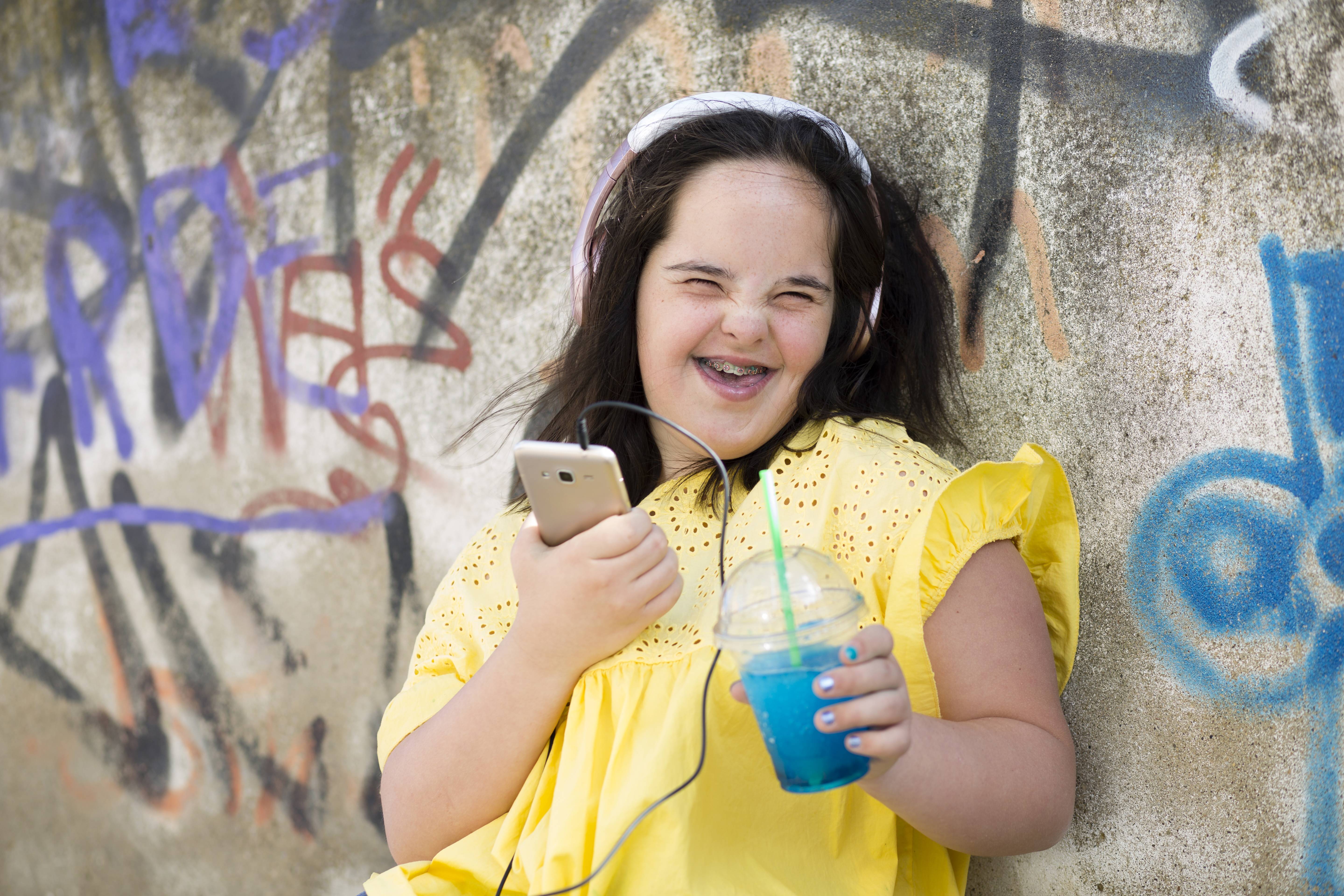 Junge Frau mit Downsyndrom mit ihrem Smartphone Junge Frau mit Downsyndrom, sie lacht in die Kamera, hält ihr Smartphone in der einen Hand und einen Plastikbecher mit einem Getränk in der anderen. Sie trägt Kopfhörer.