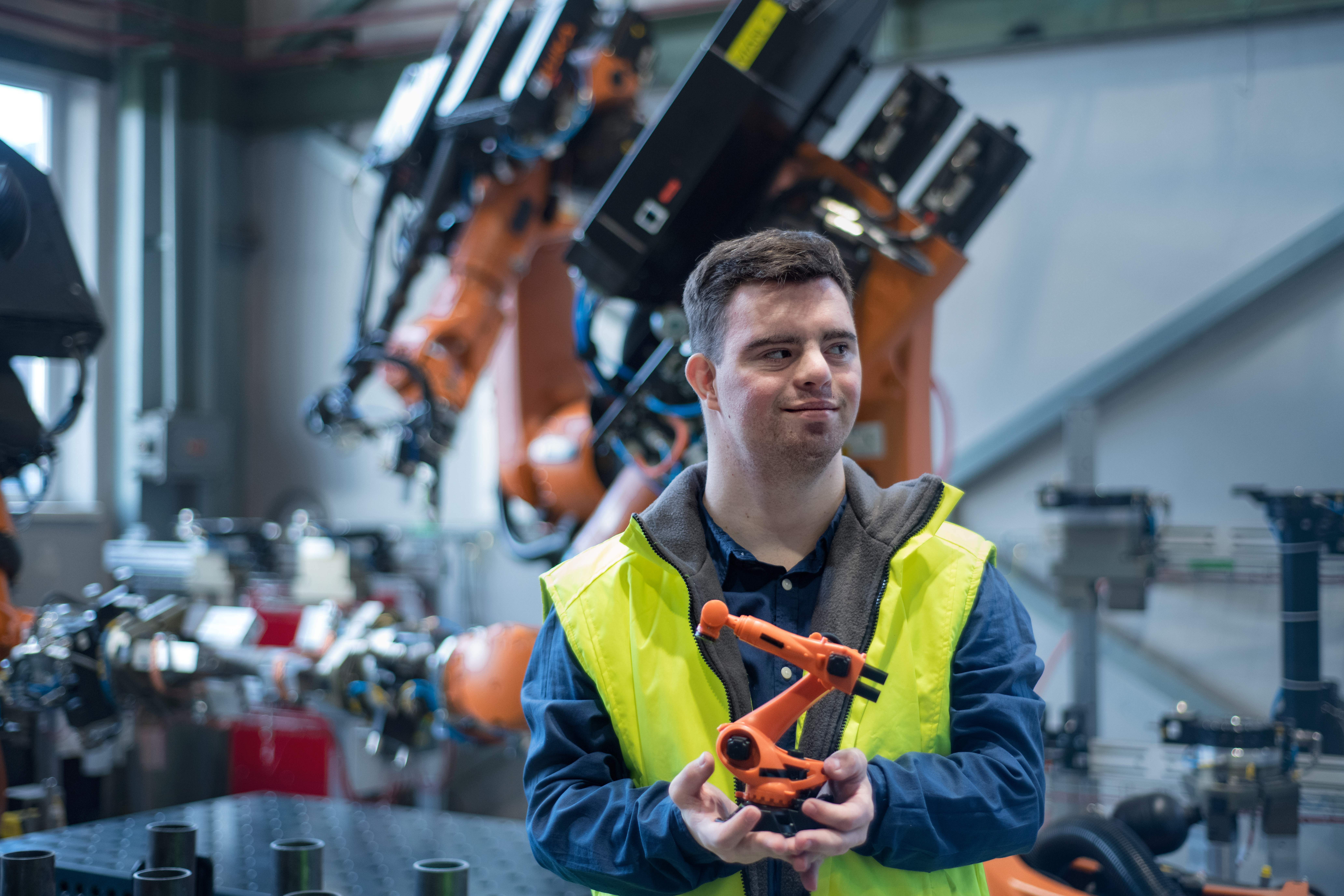 Ein junger Mann mit Downsyndrom in einer Werkshalle mit zahlreichen Maschinen, er trägt eine gelbe Sicherheitsweste