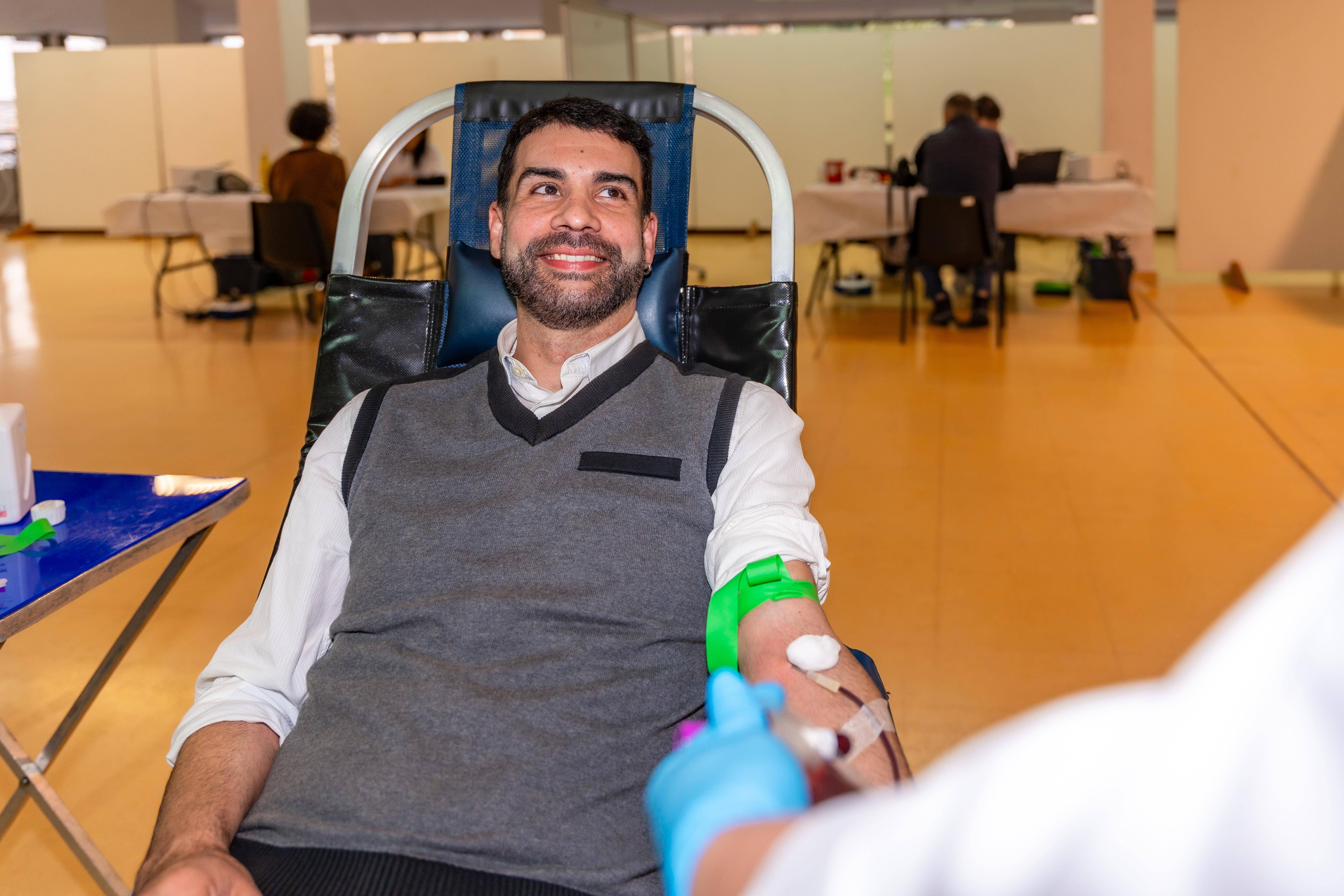 Blutspende Ein lächelnder Mann sitzt beim Blutspenden auf einem bequemen Stuhl. Im Vordergrund sieht man den Arm eines Arztes oder eine Ärztin im weißen Kittel und blauen Einweghandschuhen, die oder der sich gerade mit ihm unterhält.
