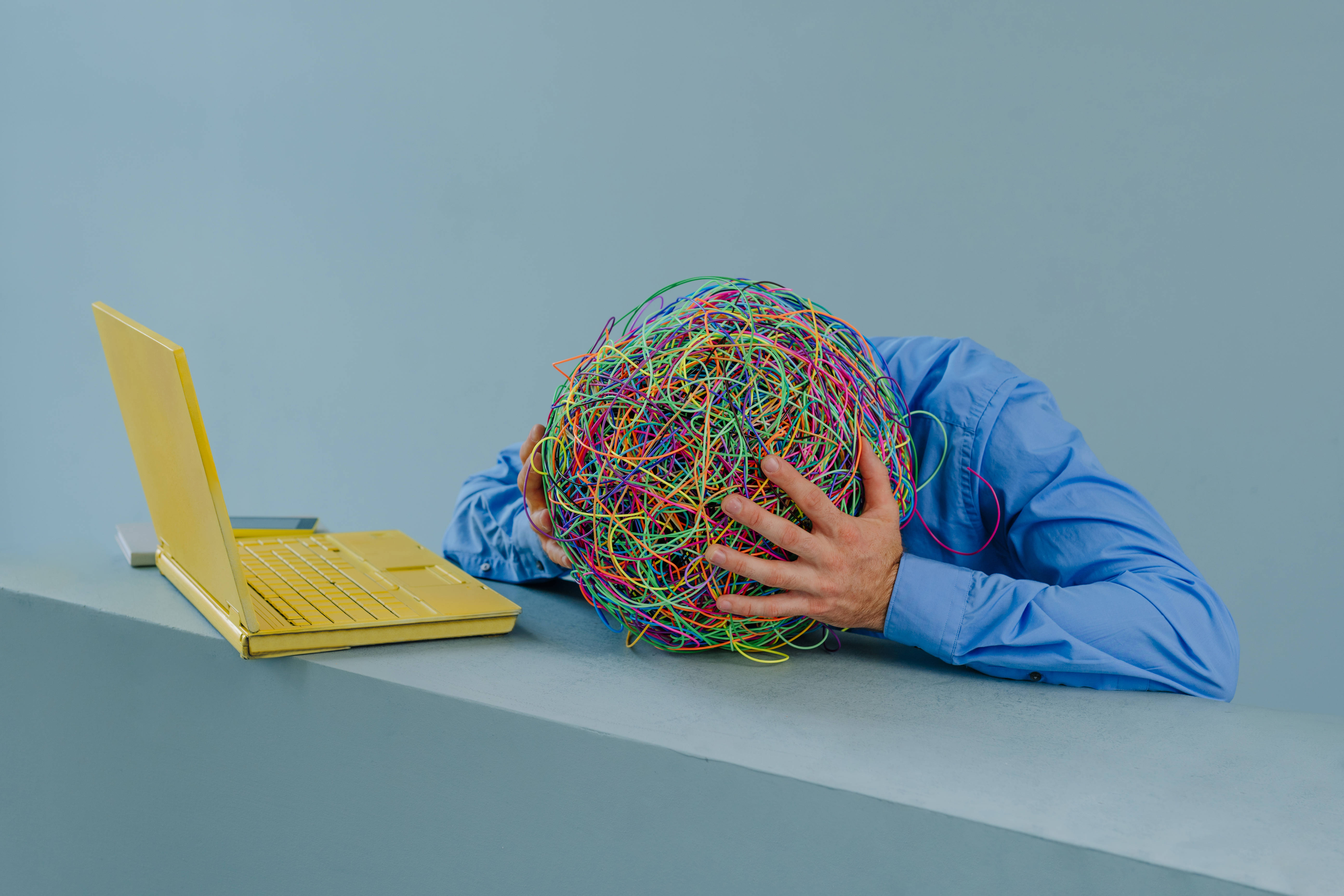 Psychische Erkrankung und Berufsleben Ein Mann im blauen Hemd sitzt vor einem gelben Notebook, er hat den Kopf auf die Tischplatte gelegt, hält sich den Kopf mit beiden Händen. Dort, wo der Kopf sein sollte, sieht man ein buntes verworrenes Knäuel als Symbol für chaotische Gedanken.