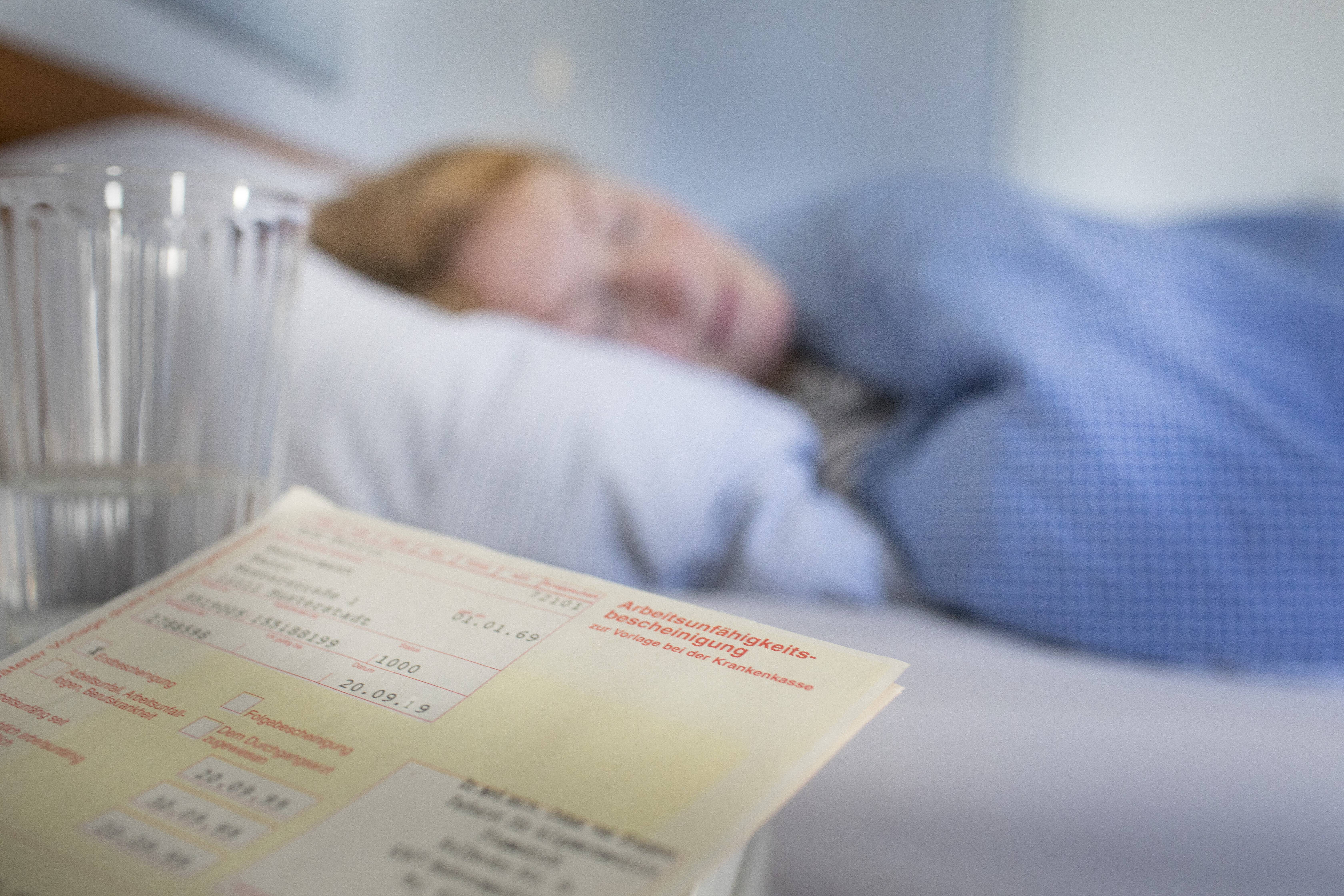 Eine Frau liegt im Bett. Auf dem Nachttisch im Vordergrund liegt eine Arbeitsunfähigkeitsbescheinigung. 