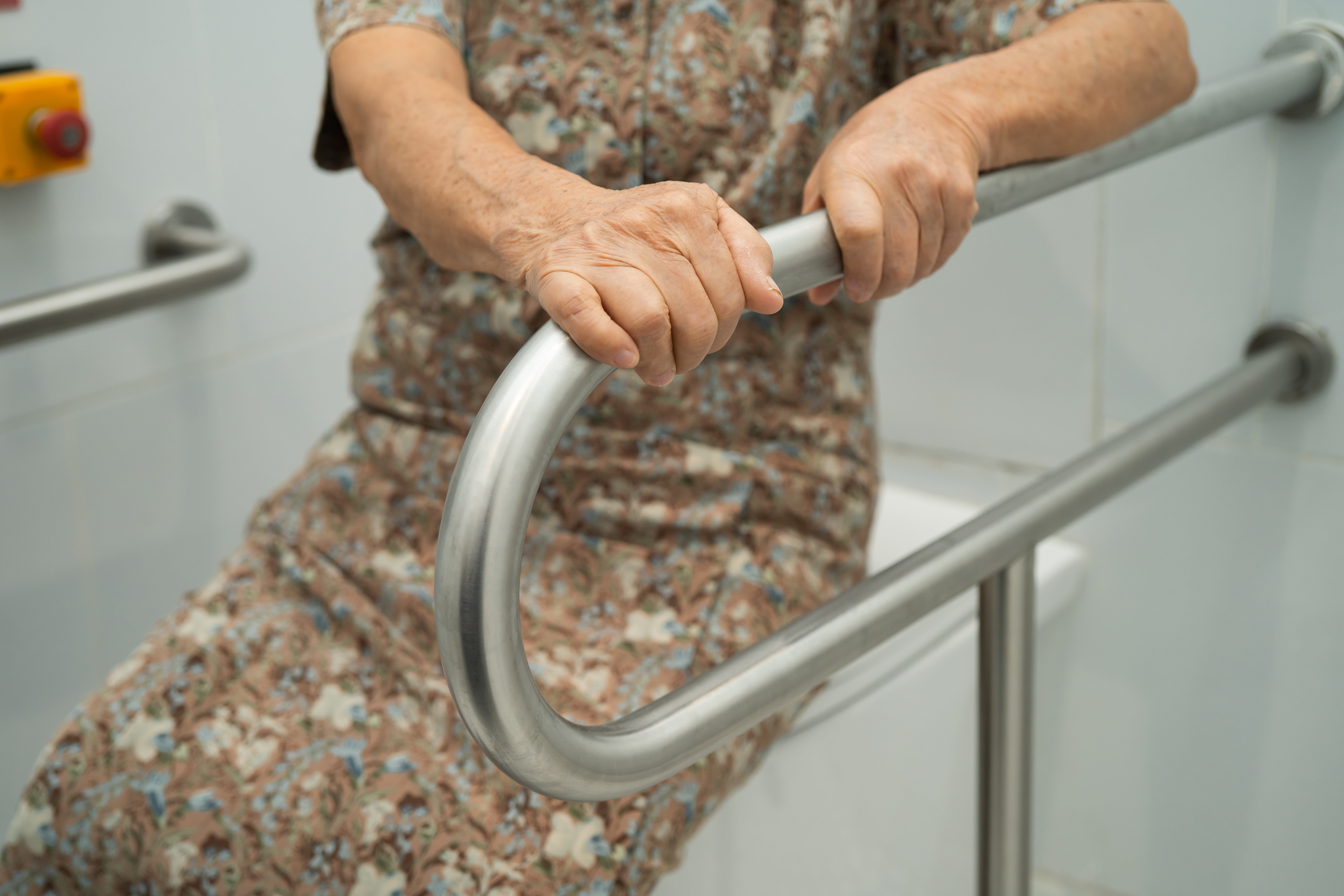 Eine &auml;ltere Frau stzt auf einem geschlossenen Toilettendeckel, sie h&auml;lt sich mit beiden H&auml;nden an einem Haltegriff fest, der neben der Toilette angebracht ist.