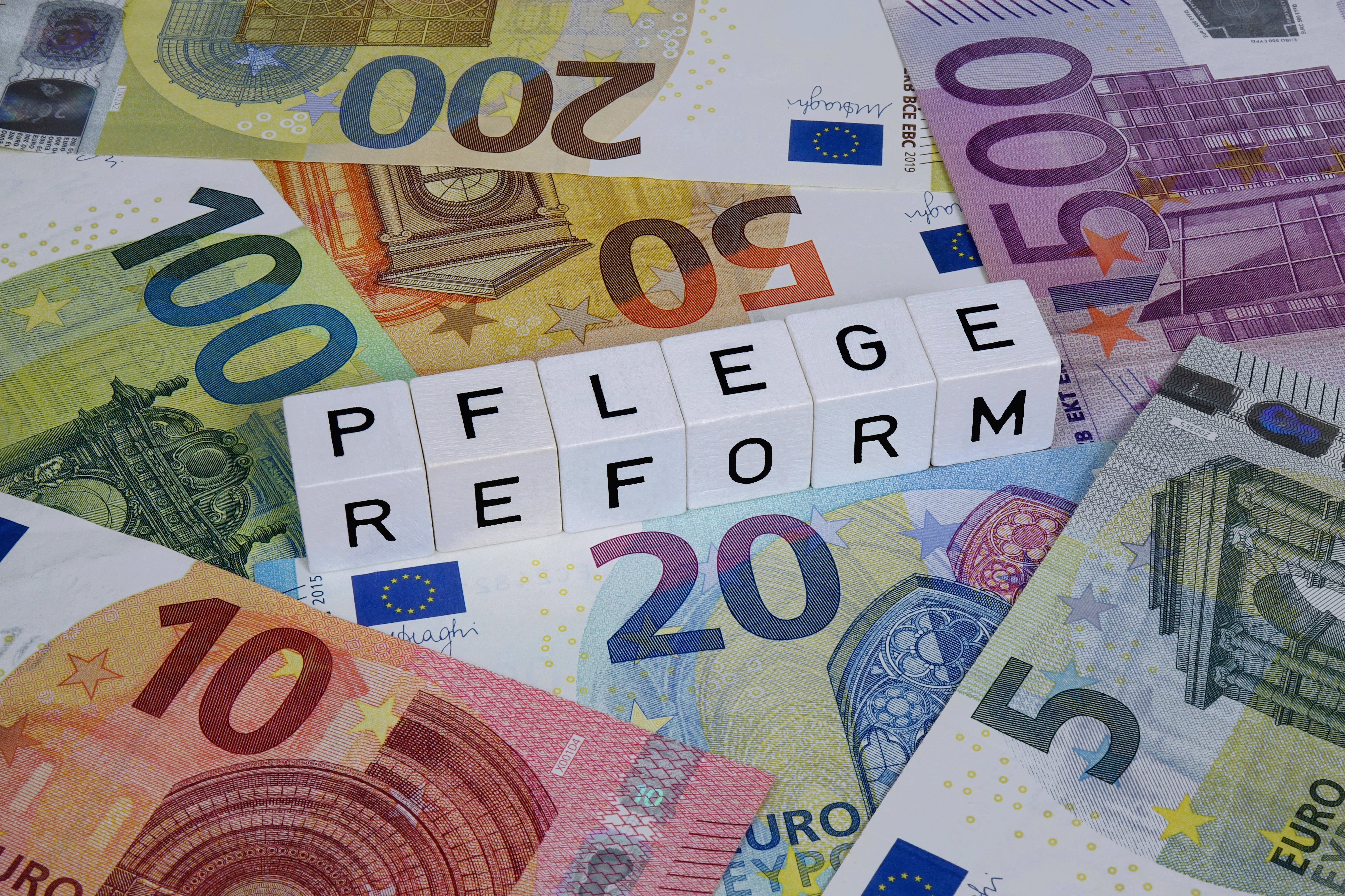 Pflegereform Geldscheine auf einem Haufen, darauf liegen Buchstabenwürfel, die das Wort "Pflegereforn" bilden