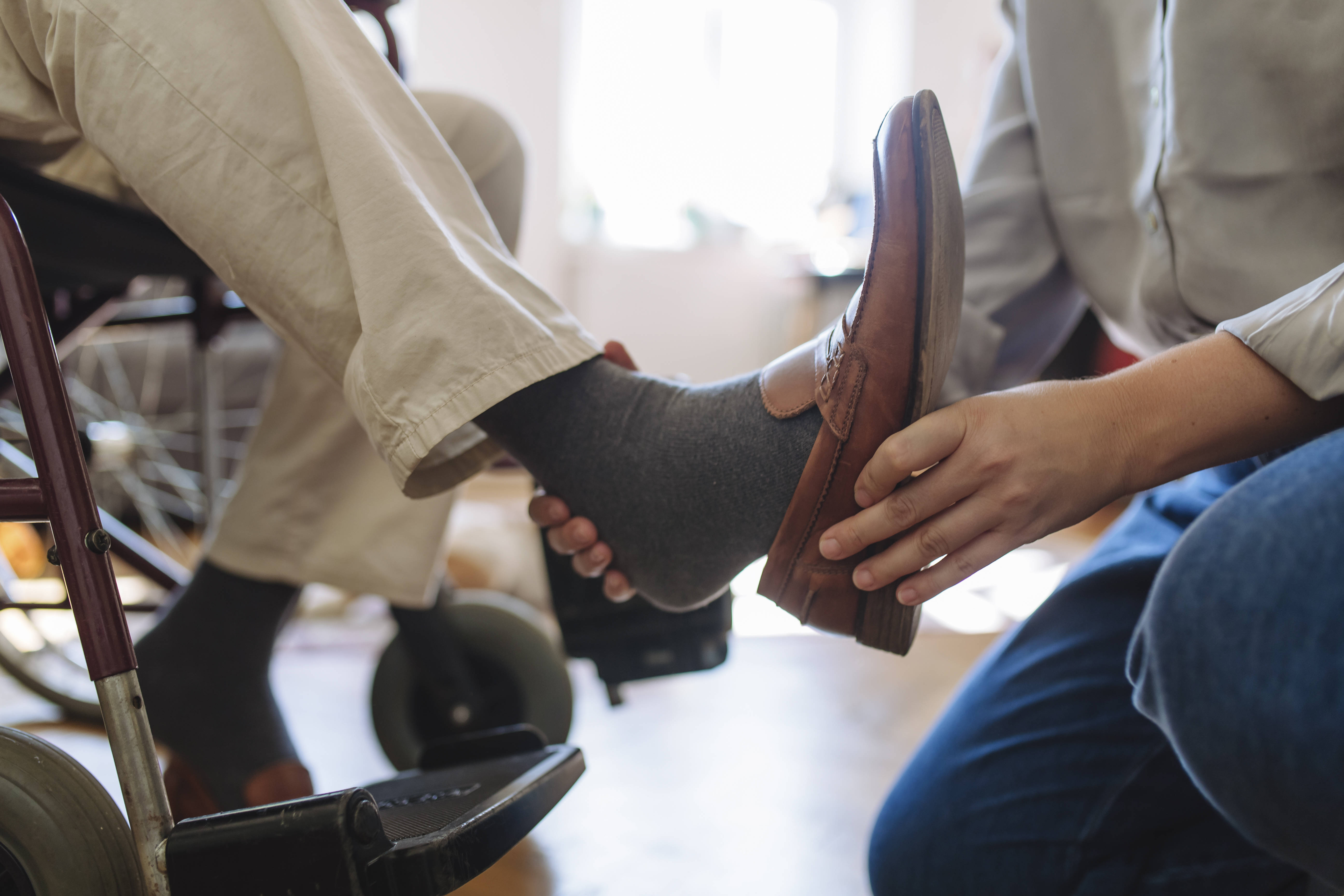 Ein Mann sitzt im Rollstuhl, eine Frau zieht ihm seinen rechten Schuh an.