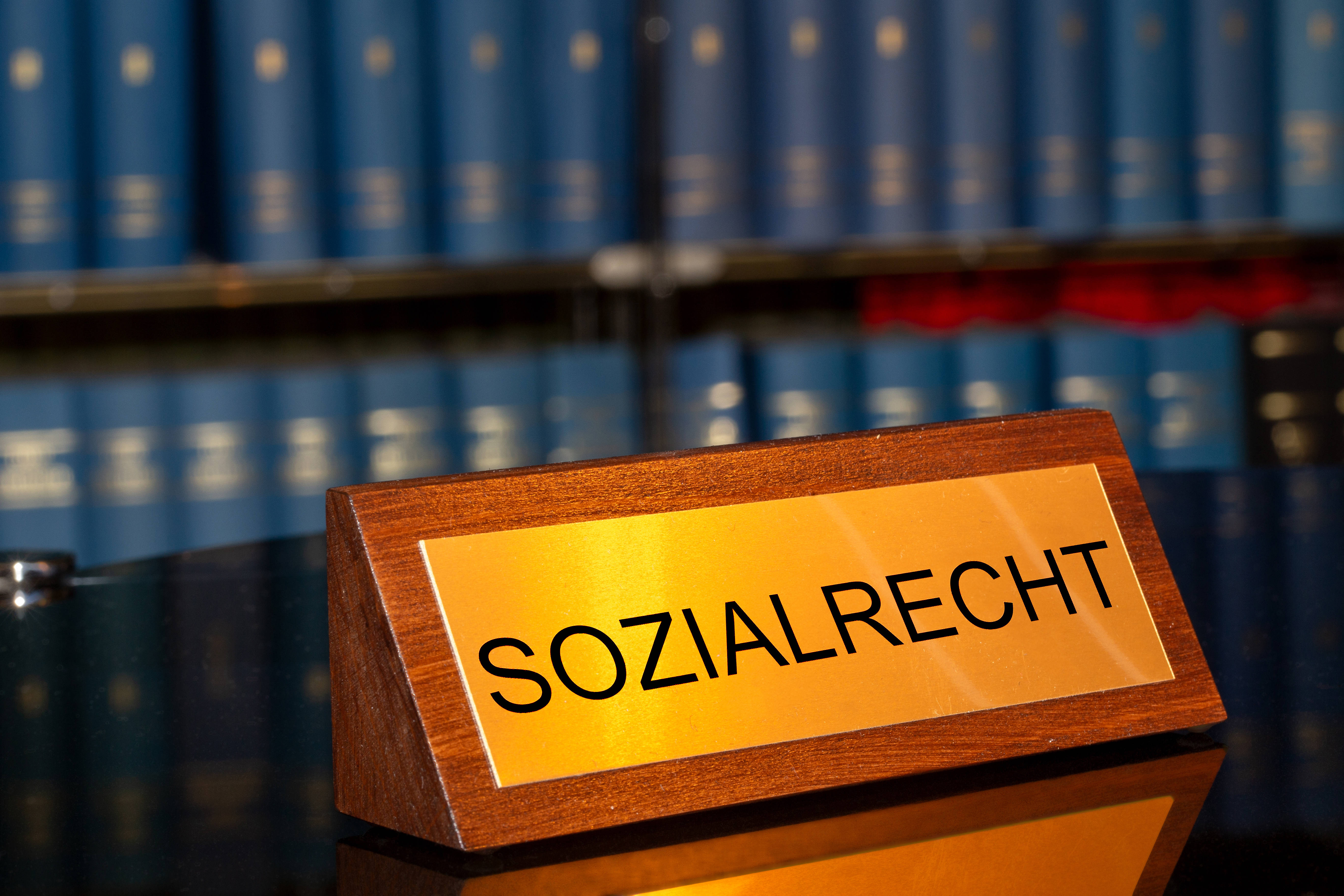 Sozialrecht Eine Bibliothek, auf einem Tisch steht ein Schild mit der Gravur "Sozialrecht", im Hintergrund sieht man viele Gesetzesbücher in einem Regal stehen.