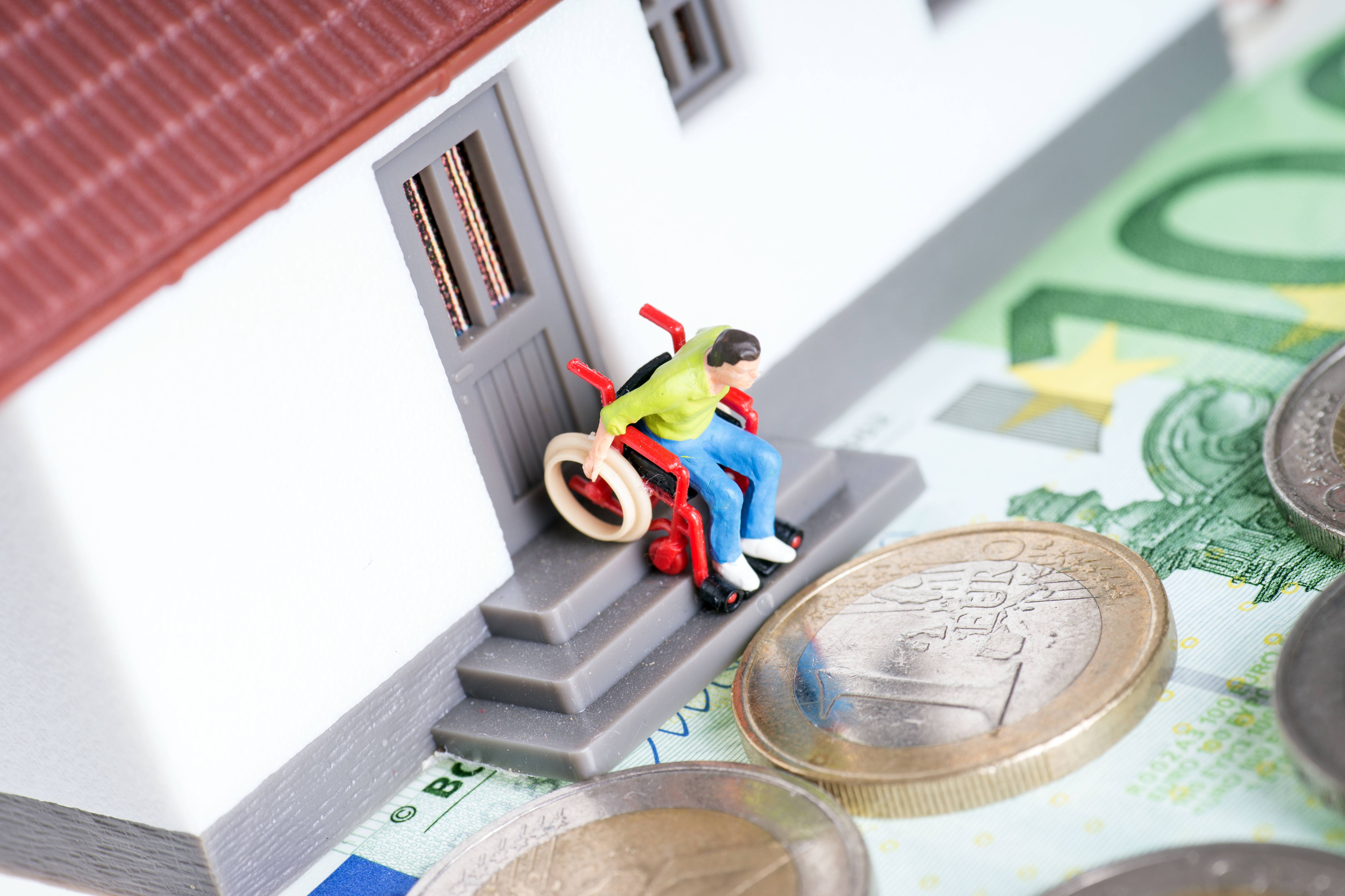 Ein Spielzeughaus, auf der T&uuml;rschwelle sind Stufen. Ein Fig&uuml;rchen im Rollstuhl steht an der Schwelle, schaut nach unten. Vor dem Spielzeughaus liegen Geldscheine und -m&uuml;nzen.