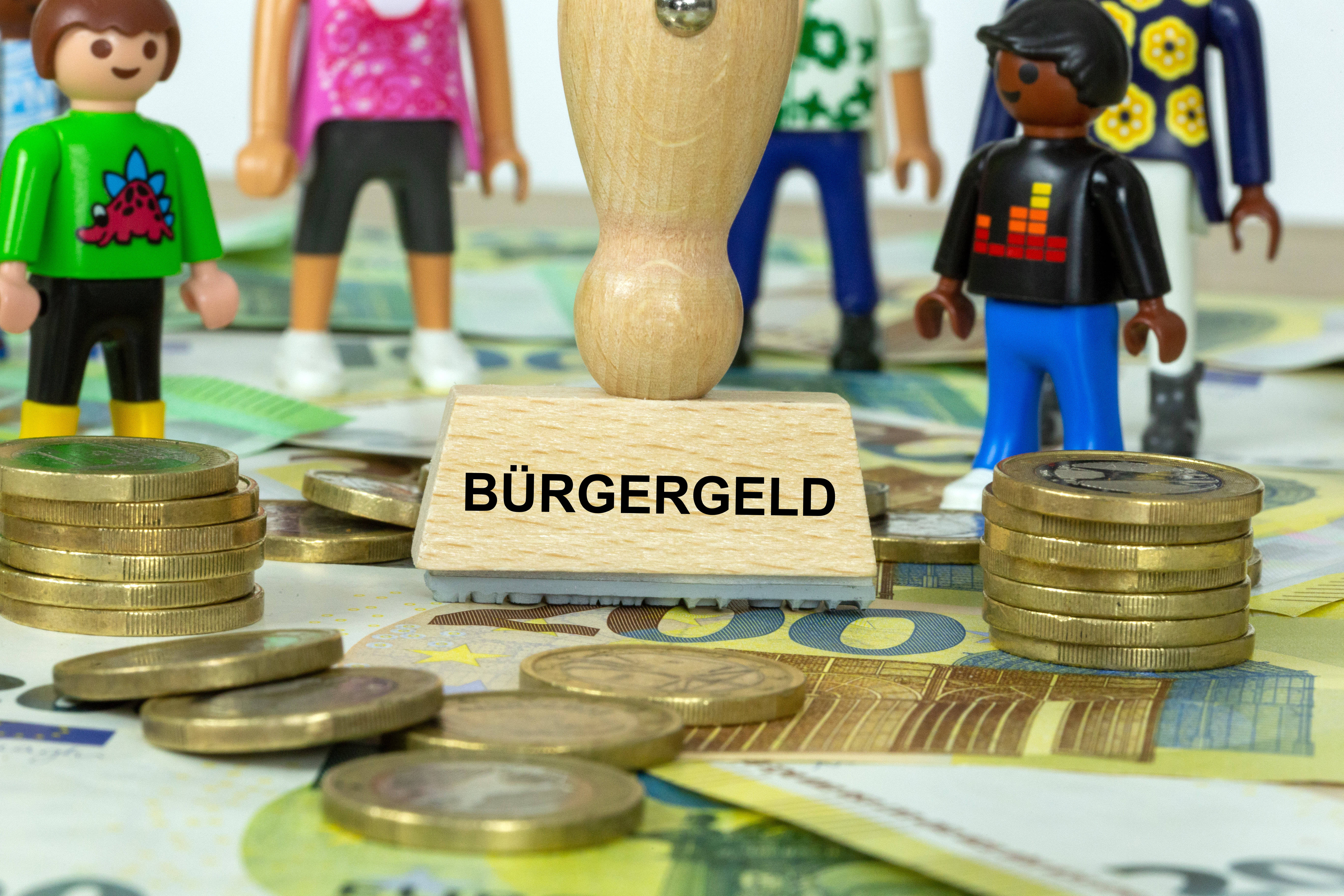 Symbolbild: Stempel mit der Aufschrift Bürgergeld und Spielfiguren auf Euro-Banknotenoten